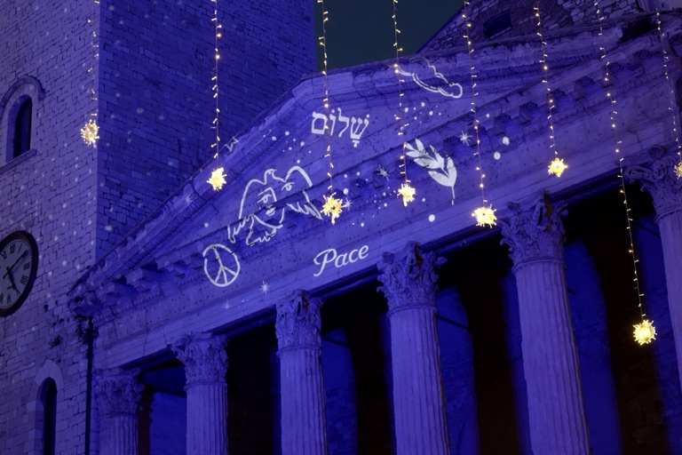 Il Tempio di Minerva in Piazza del Comune illuminato con la scritta «Pace» proiettata sul frontone